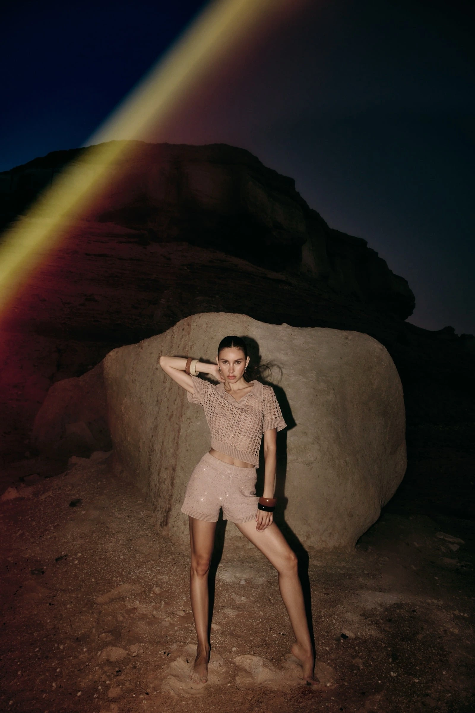 Model in a knit set standing before a vast moonlit rock at night, a rainbow lens flare rising at the edge of the sky